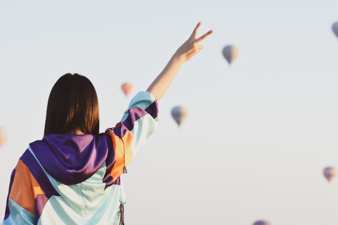 Back view of young woman in colorful hoodie raising her hand under sky with flying parachutes