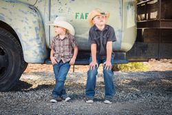 Two Boys Wearing Cowboy Hats Leaning Against an Antique Truck in a Rustic Setting