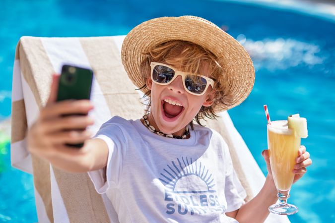 Cheerful boy holding drink and using smartphone on sunbed