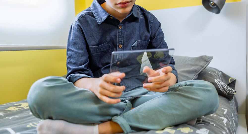 Unrecognizable boy using futuristic transparent tablet sitting over his bed