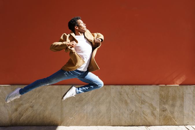 Black male in camel coat leaping against am outdoor wall