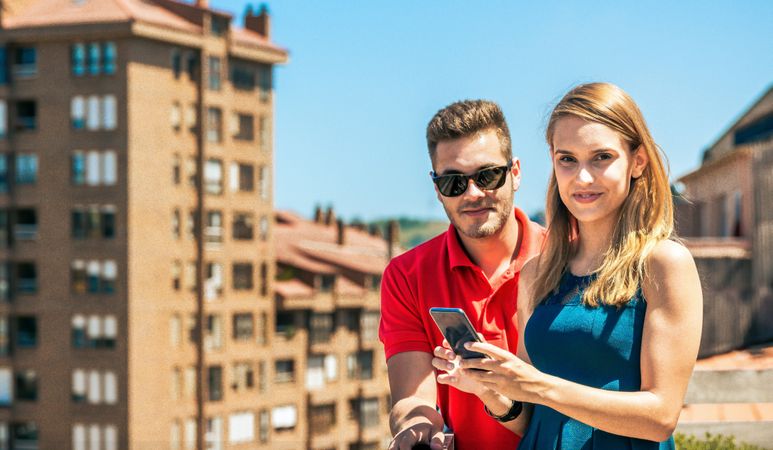 Young couple with mobile looking at camera in rooftop on a summer day