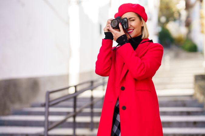 Female photographer in stylish clothes taking photo while standing on stairs in city