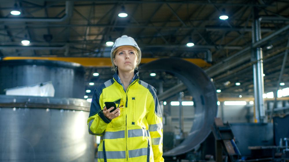 Female Industrial Worker in the Hard Hat Uses Mobile Phone While Walking Through Heavy Industry Manufacturing Factory.