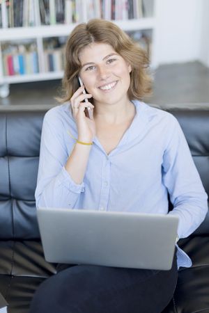 Smiling woman sitting on sofa at home using a laptop and speaking on phone