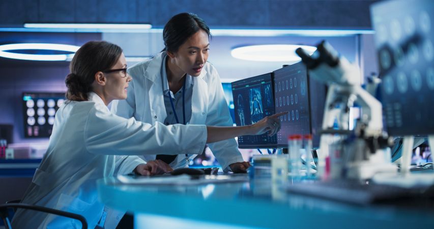 Medical Hospital Research Laboratory: White And Asian Female Scientists Talking And Using Desktop Computer With CT Scans Of Human Brain.