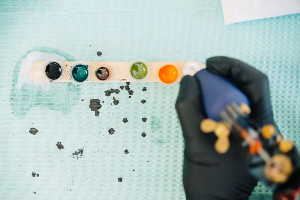 Top view of tattoo gun being loaded from a row of ink pots on blue paper