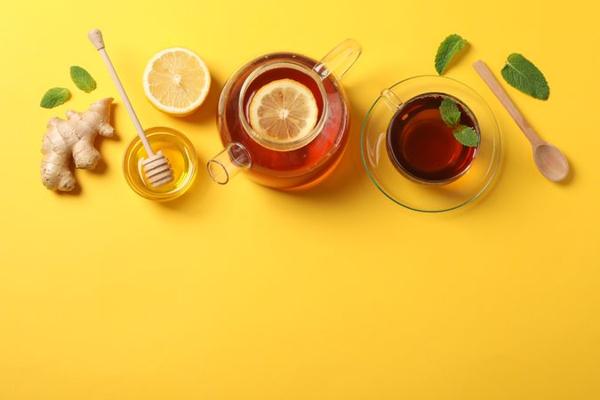 Flat lay. Cup of tea, teapot, lemon, ginger, mint, honey and dipper on yellow background, copy space