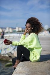 Smiling female with curly hair in bright green shirt sitting listening to music on pier