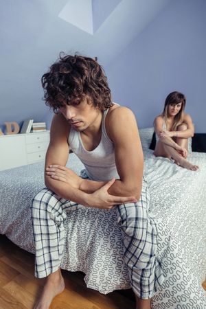 Angry man sitting on edge of bed with woman in background