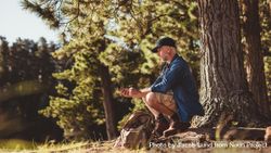 Mature man hiker sitting by a tree in forest and checking his position by using a compass bxq3d0