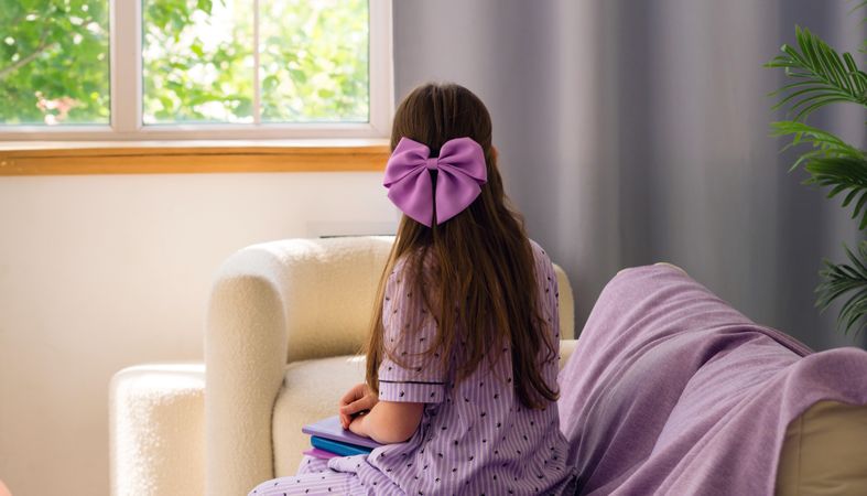 Young girl looking out window, wearing purple bow.