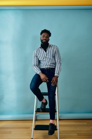 Black man in blue studio resting on step ladder