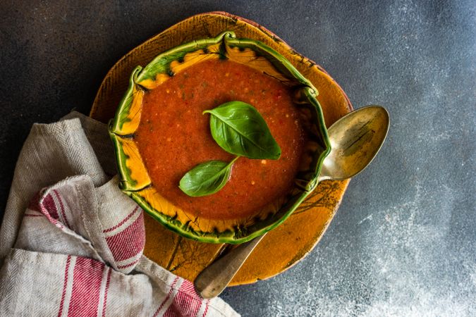 Green and orange ceramic bowl of gazpacho soup with basil leaves and kitchen towel