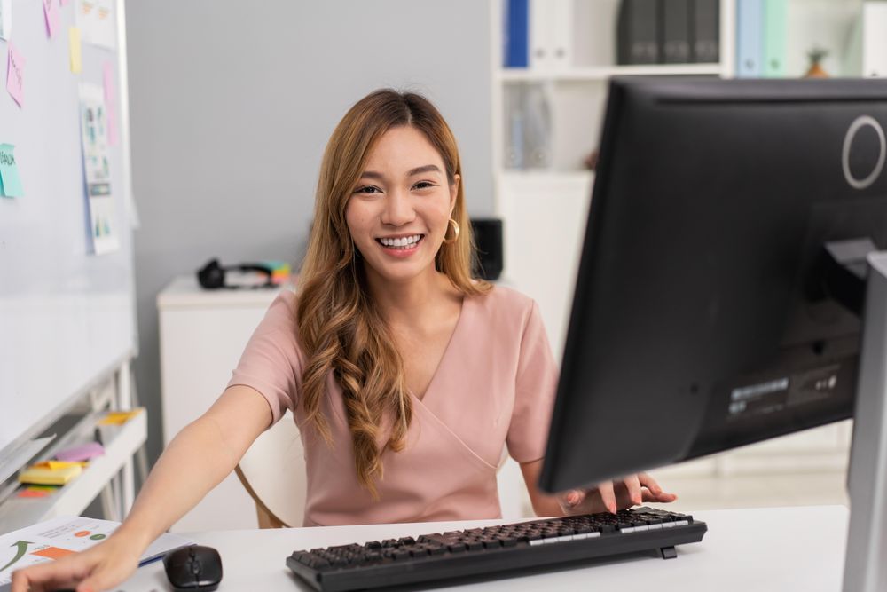 Happy Asian woman sitting in office behind computer and monitor