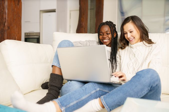 Two women in jeans and sweater watching something together on a sofa on a pc