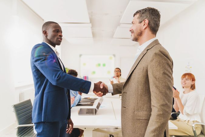 A Black man and a white man shaking hands in a modern office while colleagues applaud in the background