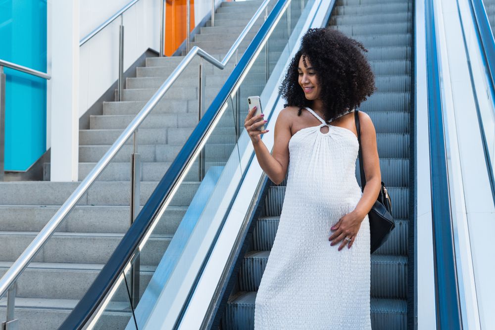 Pregnant Black woman browsing smartphone on escalator