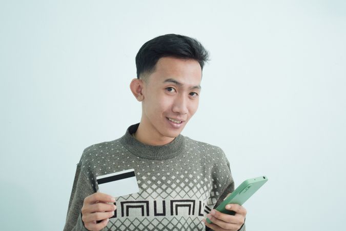 Curious Asian businessman in suit holding blank credit card and smartphone