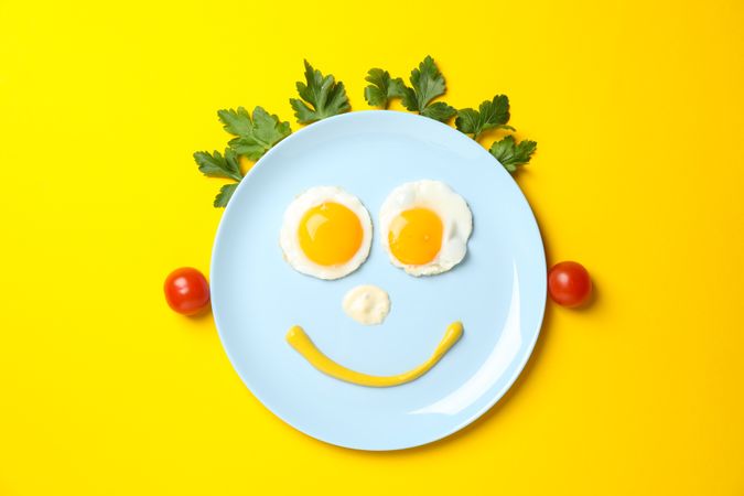 Looking down at blue plate with smiley face on it made of eggs and condiments, with vegetable hair