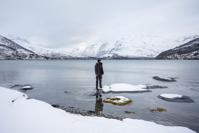 Solitary tourist on fjord shore against snowy hills in cold overcast weather