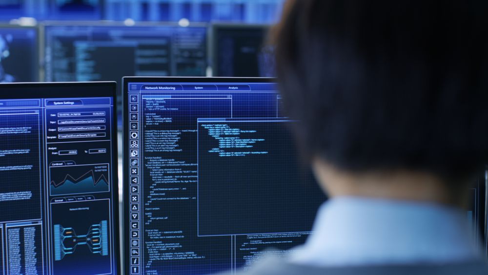 In the System Control Room Technical Operator Stands and Monitors Various Activities Showing on Multiple Displays with Graphics.