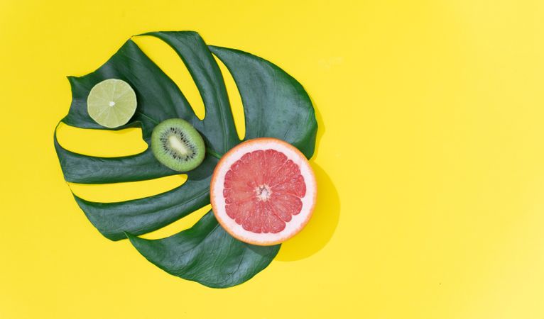 Monstera leaf with red grapefruit, lime and kiwi on bright yellow background