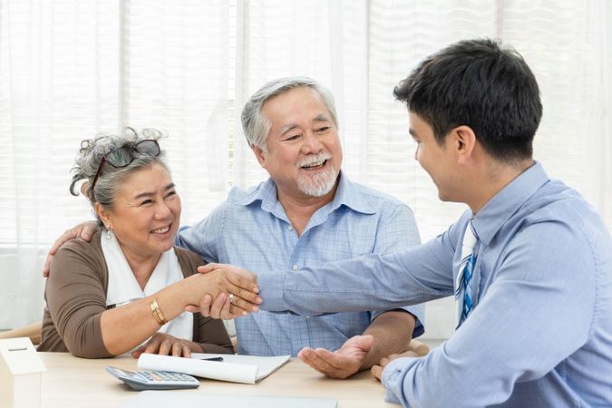Smiling Asian couple making purchase with mature woman shaking hands with real estate agent