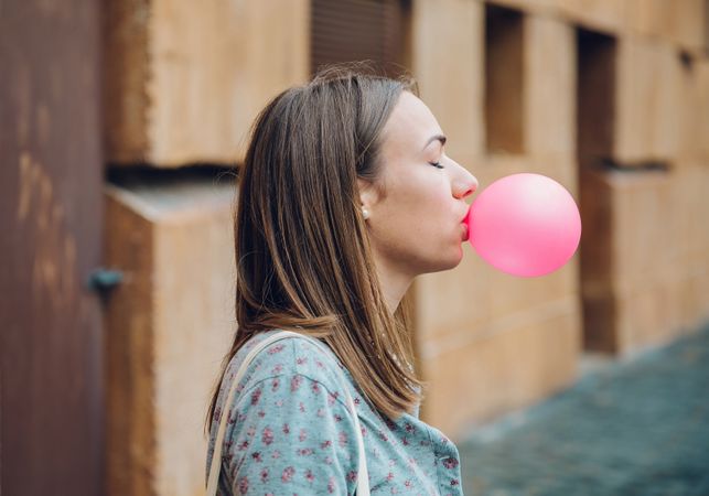 Side view of female standing outside in front of stone wall blowing bubble with pink gum with eyes closed