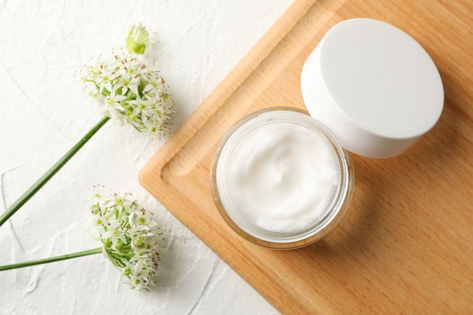 Jar with cream, allium flowers and board on blank background, top view