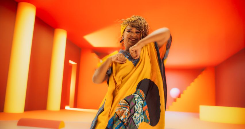 Black Woman in African Outfit Dancing Energetically in Geometric Abstract Orange Environment. Creative Female Performing Dance Choreography in Bright Studio, Making Moves, Practicing.