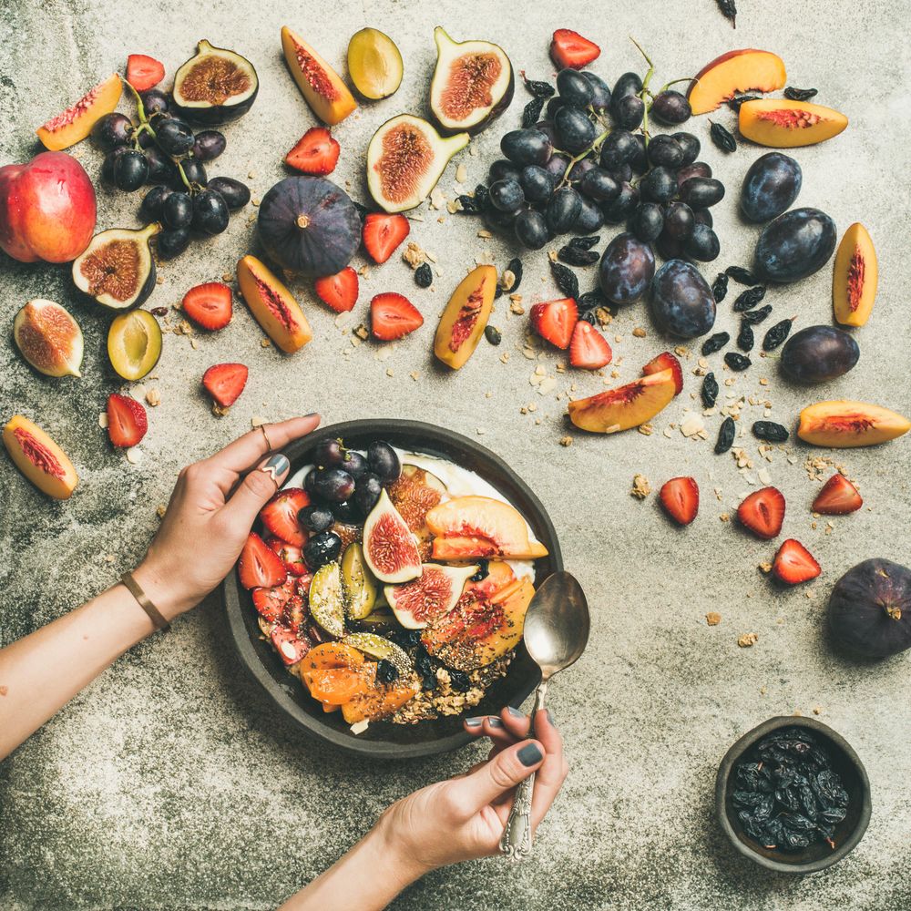 Woman holding bowl of yogurt with spoon over table of figs, peaches, and chia seeds, square crop