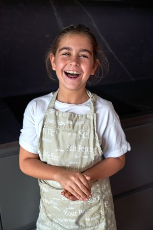 Cheerful little girl in apron standing in kitchen