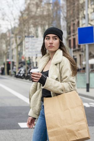Portrait of young woman walking on a cold city street, with takeaway coffee and shopping bag