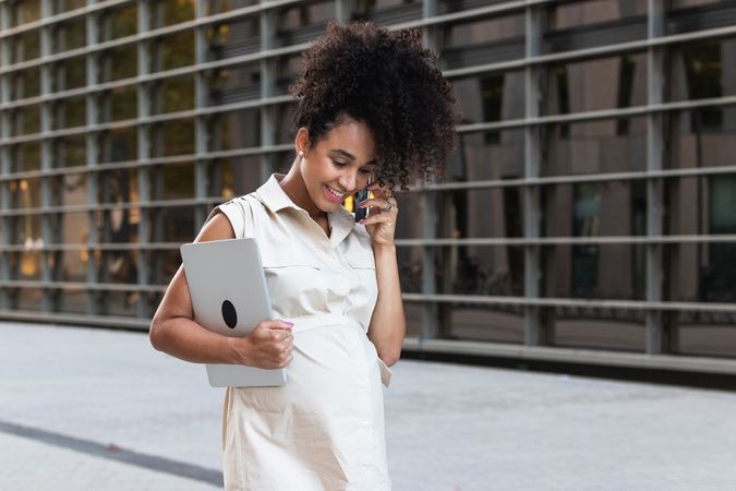 Smiling Black pregnant woman talking on smartphone and carrying a laptop
