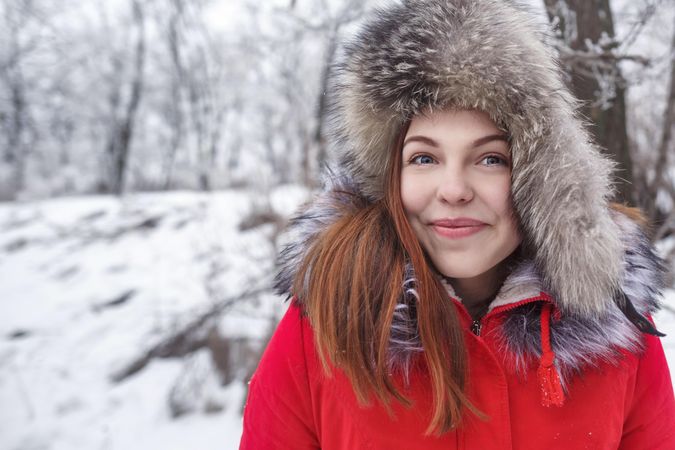 Smiling young woman in red winter coat, fur hat in snowy forest
