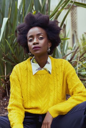 Woman with afro hair wearing yellow knit sweater sitting beside green plant