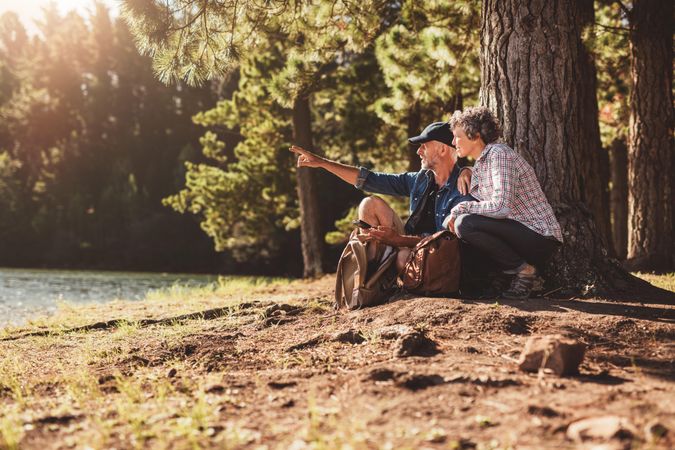 Mature couple sitting by a tree in forest with man showing something to woman