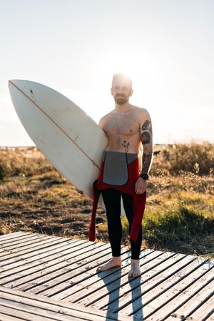 Tattooed man in wetsuit standing on boardwalk leading to the beach with surfboard