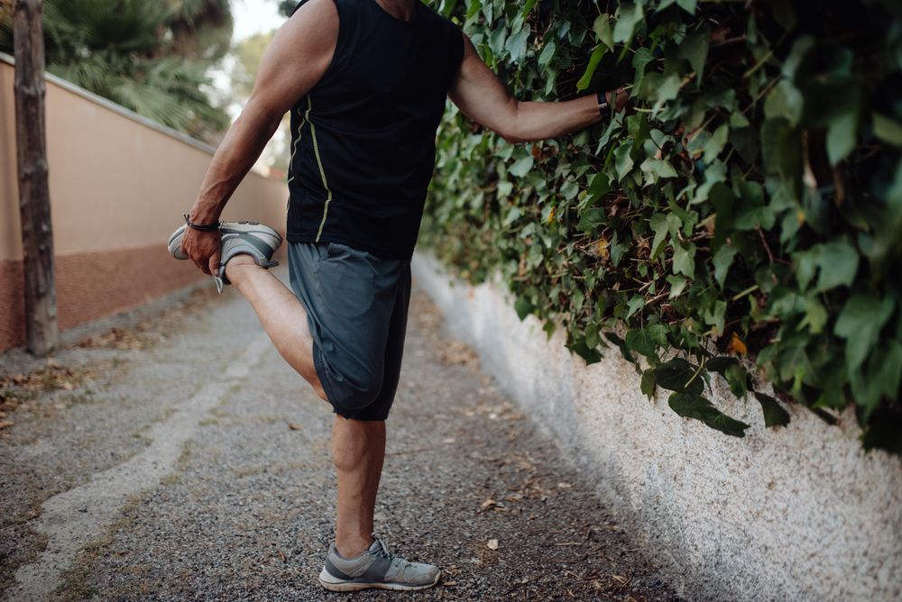 Older man stretching leg holding botanical wall in street