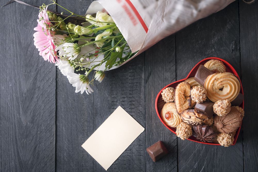 Top view of heart-shaped box with sweets, bouquet of flowers and blank message card on rustic table