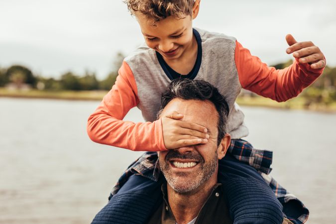 Playful young boy covering his fathers eyes while sitting on his shoulders
