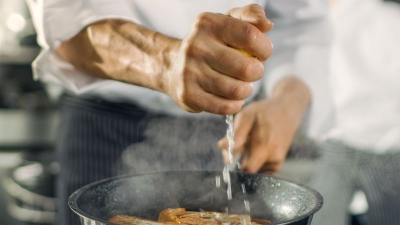 Professional Chef Squeezes Lemon Juice onto Hot Pan with Red Fish Fillet on it. He Works in a Modern Kitchen.