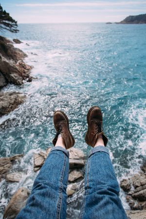 Hiker sitting on rocks overlooking ocean in leather boots, vertical