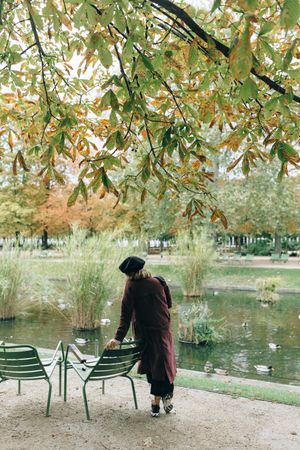 Back view of woman in red coat with dark hat standing beside river under a tree in Paris, France