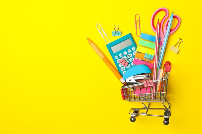 Shopping trolley with school supplies on yellow background, space for text