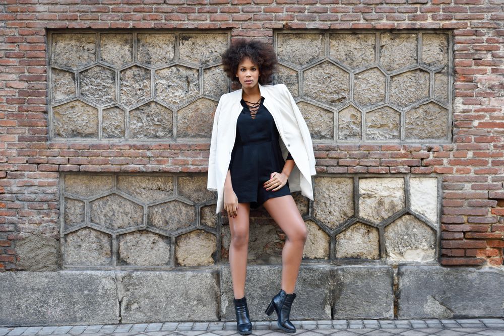 Female in short dress, heels and blazer leaning on brick wall and looking at camera