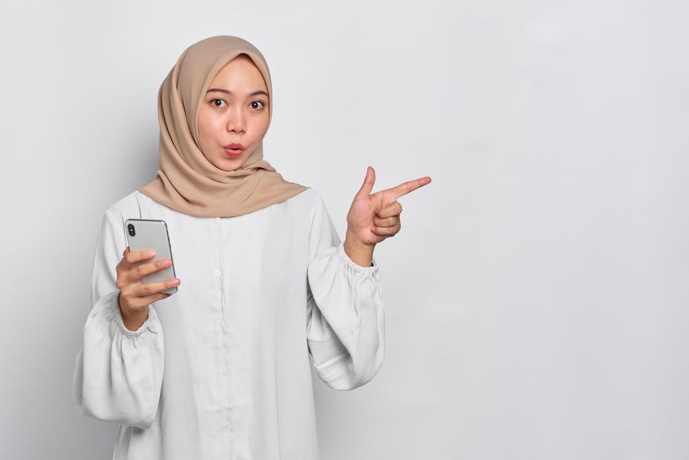 Surprised Asian female in headscarf pointing to the side while holding cell phone