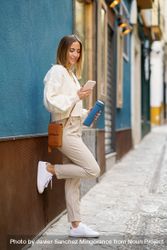 Happy woman in cream leaning on wall outside and checking phone 4d8B7L