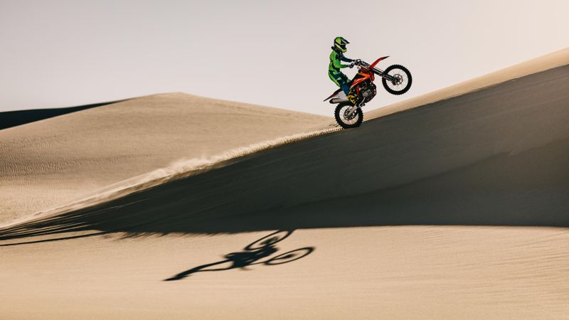 Side view of motocross bike rider riding up over sand dunes
